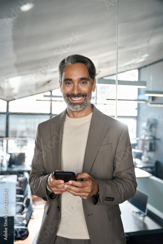 Happy mature 45 years old business man executive using smartphone standing in office. Smiling middle aged businessman ceo leader entrepreneur holding mobile cell phone at work. Vertical portrait.