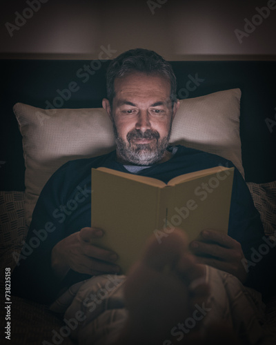 Man alone in pajamas reading a book lying on bed at late at night. Boy sitting in bedroom reads a novel at home. Male enjoy free time at home in relax.  Lifestyle, bedtime, rest and people concept