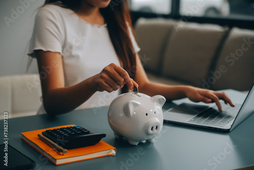 Young Asian woman saving for retirement Saving money through a piggy bank and taking notes on notebook, savings concept.