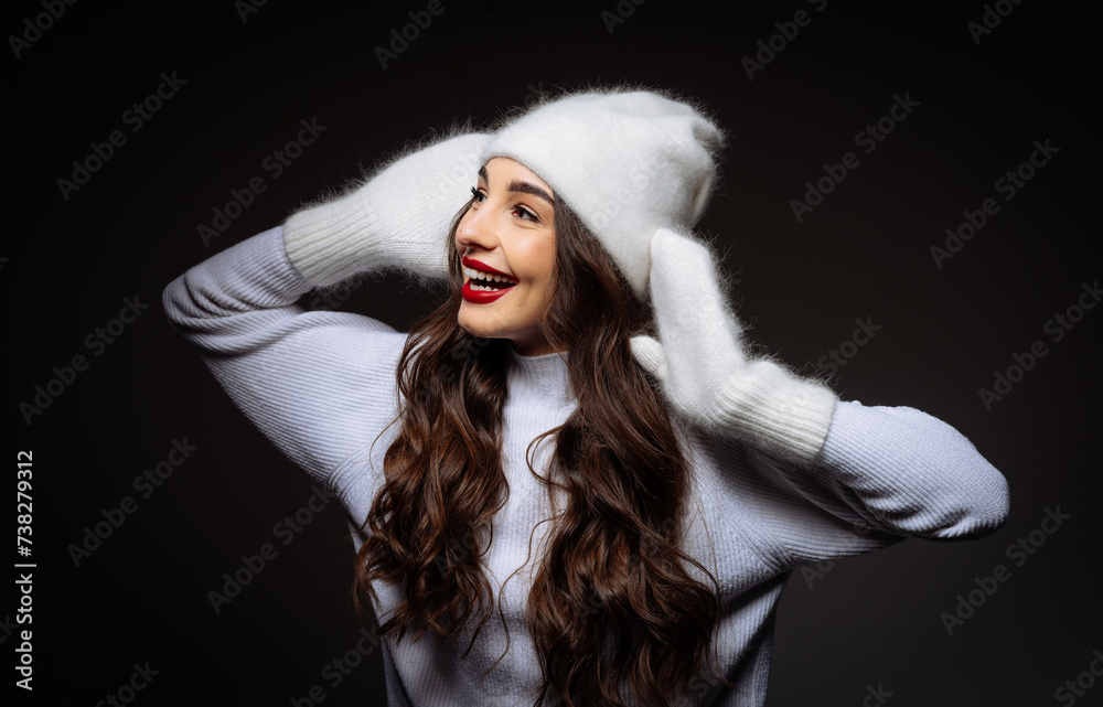 Beautiful young woman wearing winter hat is smiling while standing against black background in the studio Beautiful young woman wearing winter hat is smiling while standing against black background in the studio