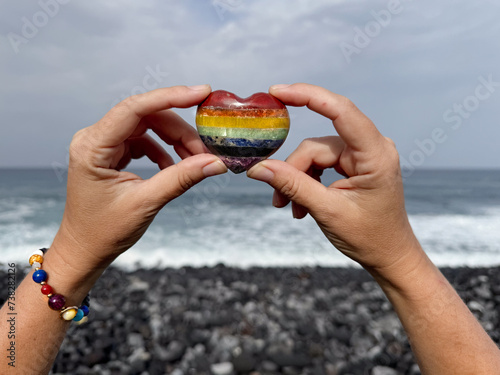 Hands holding rainbow colored stone heart on ocean shore, meditation, spiritual journey, connection with Nature photo