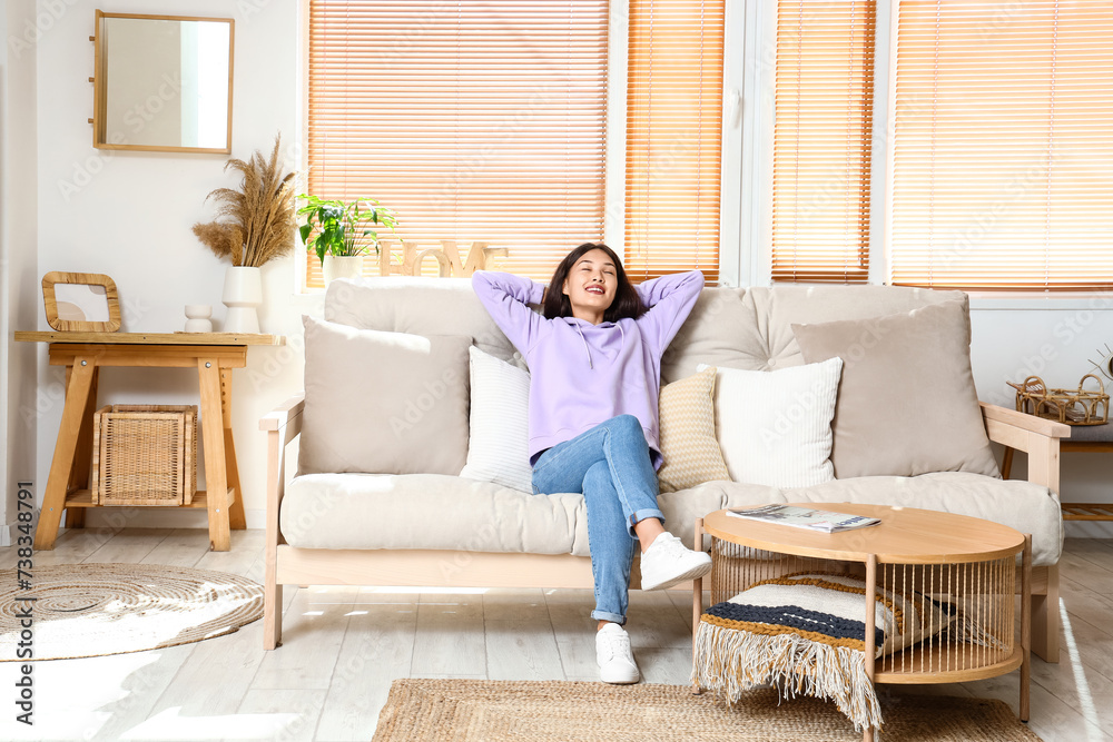 Beautiful young Asian woman in stylish hoodie sitting on sofa at home Beautiful young Asian woman in stylish hoodie sitting on sofa at home