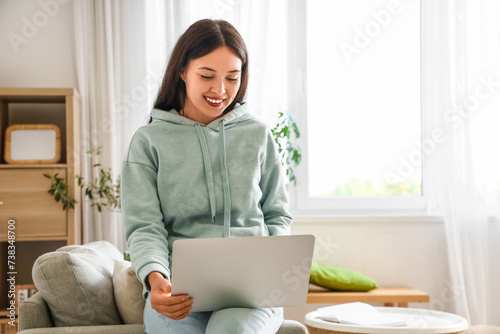 Beautiful young Asian woman in stylish hoodie with laptop at home © Pixel-Shot