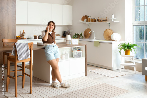 Pretty young woman talking by phone in modern kitchen