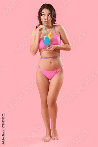 Sexy young woman in swimsuit and with Blue Lagoon cocktail on pink background