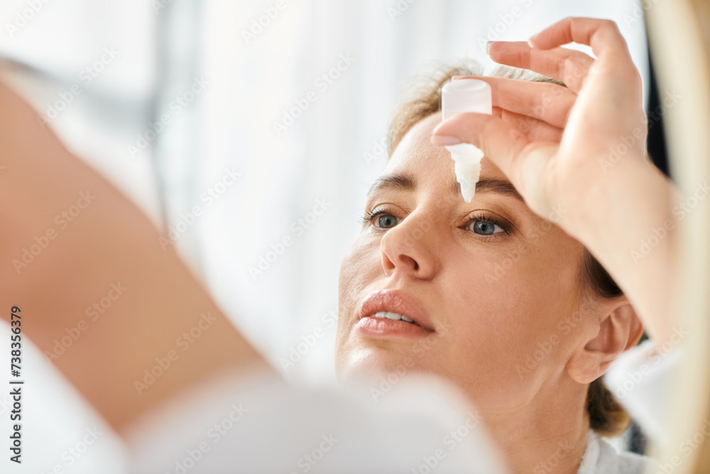 Naklejka premium beautiful blonde woman with collected hair in bathrobe putting in eye drops in her bathroom