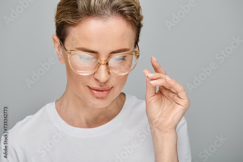appealing blonde woman in casual attire with collected hair posing with her glasses on gray backdrop
