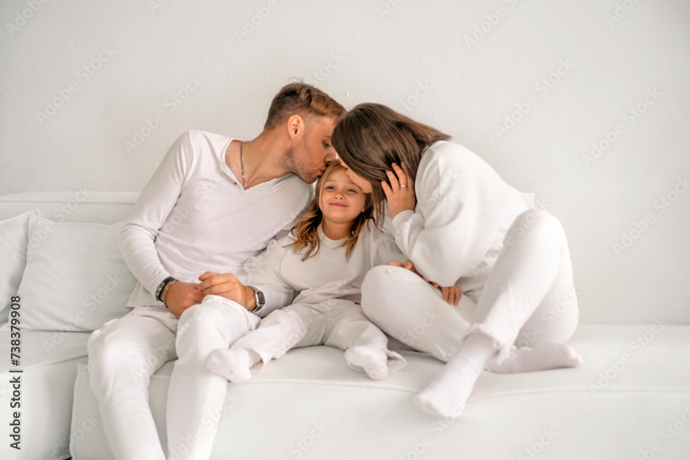 A young family with their daughter are sitting on the couch, kissing the child on both sides. Loving parents A young family with their daughter are sitting on the couch, kissing the child on both sides. Loving parents