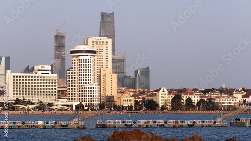 Coastline scenery of Qingdao, Shandong, China, with modern high-rise buildings in Huiquan Bay in the distance and sea water and rocks nearby