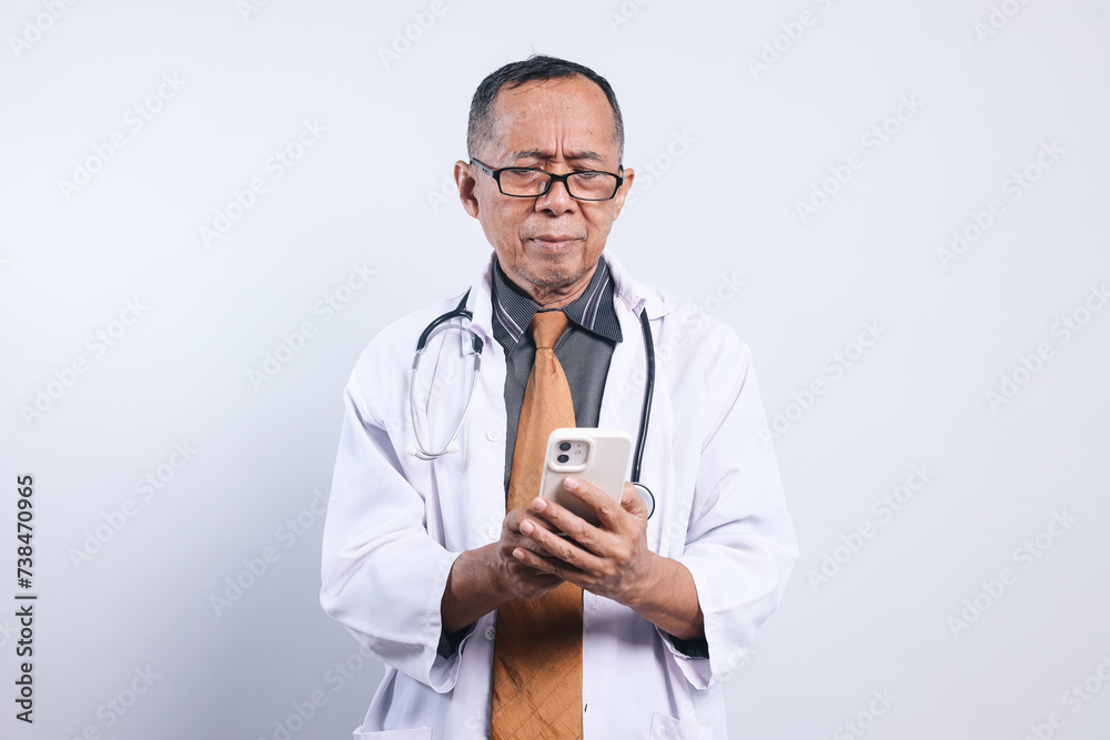 Asian senior doctor with stethoscope and white coat looking at phone with confused expression 