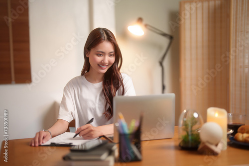 Young asian women reading financial business report on laptop and taking notes in paper while working and analysis about marketing strategy of new startup project in living room at home office