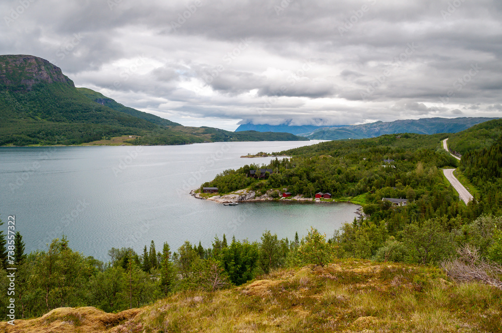 The coast of a fjord in Norway in a mountainous landscape with forests, a road and small wooden houses over the water.