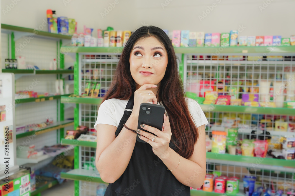 Thoughtful Asian people holding her phone, cashier is wearing black apron standing in a groceries / convenient store