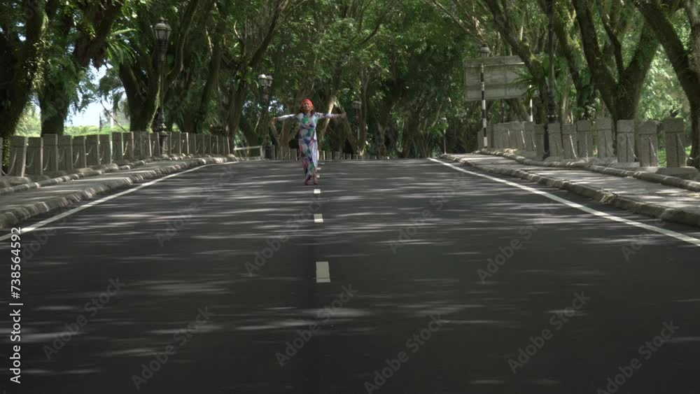 Asian woman in a colored stylish dress with turban walks and dance along the asphalt road under shady trees in the park. Portrait of a woman in motion between trees. Asian woman in a colored stylish dress with turban walks and dance along the asphalt road under shady trees in the park. Portrait of a woman in motion between trees.