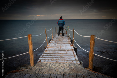 Person standing at the edge of a wooden pier in the sea at sunrise . People active outdoors enjoy ocean