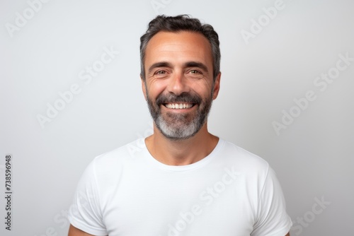 Handsome middle-aged man in white t-shirt smiling and looking at camera while standing against grey background