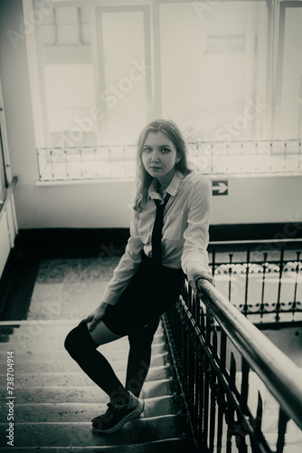 A beautiful young student, wearing a uniform, stands on the steps of an institute on a typical school day. The image is in monochrome.