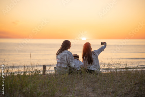 Back view of two caucasian female friends and a child making selfie at sunset on the Baltic seashore photo