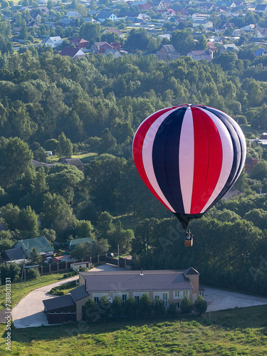 A blue, white, red hot air balloon flies over the countryside. On the ground there are small houses, fields. Summer day. Balloon flight.