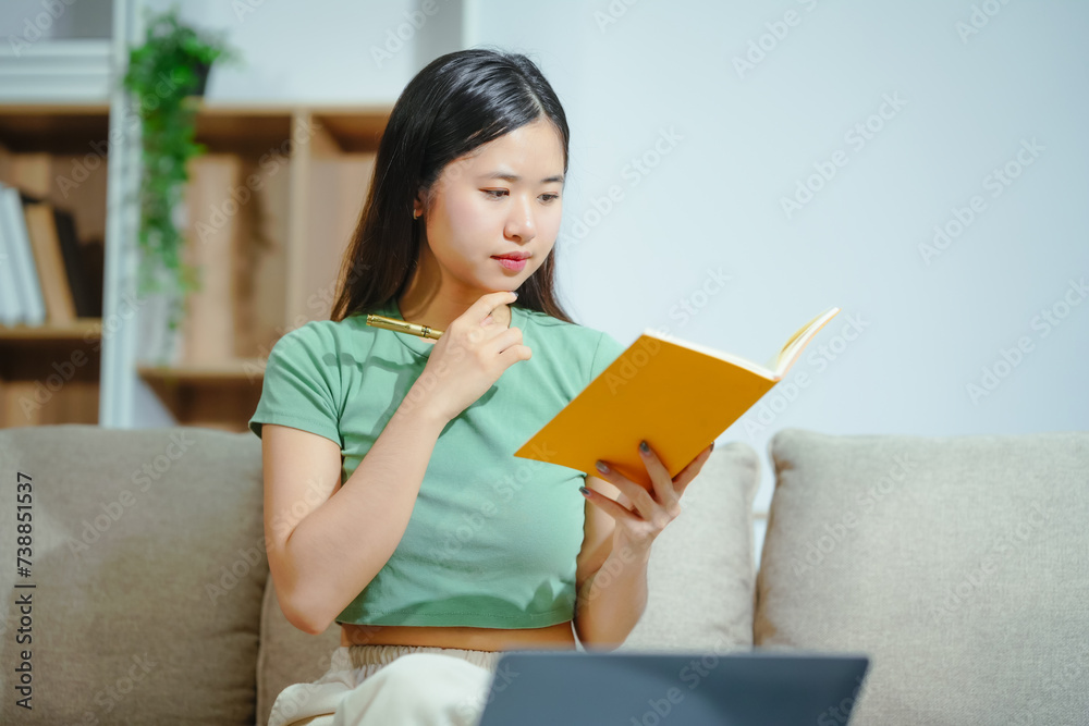 young Asian girl engages in online learning, doing homework on her sofa in the living room. video classes, combining comfort and education at home. young Asian girl engages in online learning, doing homework on her sofa in the living room. video classes, combining comfort and education at home.