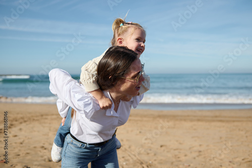 Happy young mother giving piggyback ride to her little daughter, enjoying walk together on the sandy beach