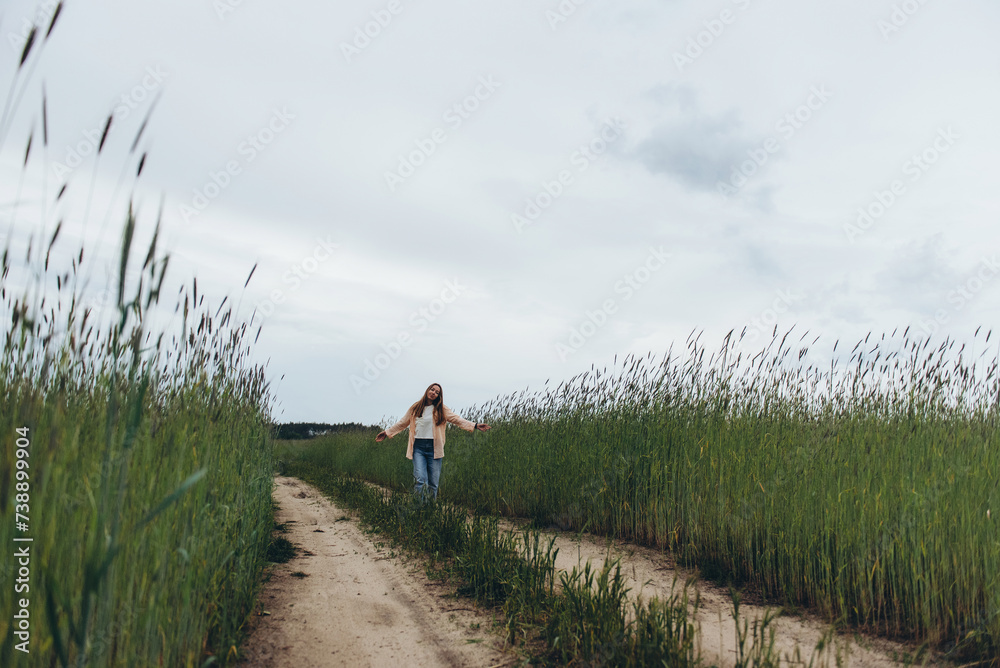 A girl, a woman stands on the road in the middle of a field of green wheat. Cool and cloudy summer day. He dreams with his eyes closed and his arms spread out to the sides like wings.