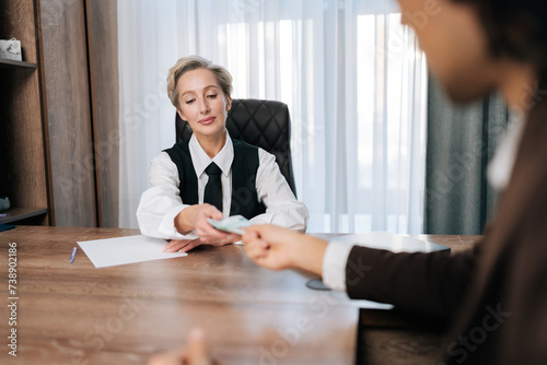 Back view of successful businessman in suit giving money to grateful professional middle-aged female partner as reward for successful business contract, sitting at office desk.