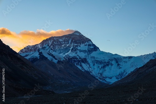 Experience the breathtaking spectacle of sunrise over Mt. Everest as its towering silhouette casts a majestic shadow against the dawn-lit sky, while vibrant crimson hues dance atop the veiling clouds.