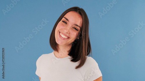 Close up, beautiful young brunette woman dressed in white top laughs looking at camera isolated on blue background in studio