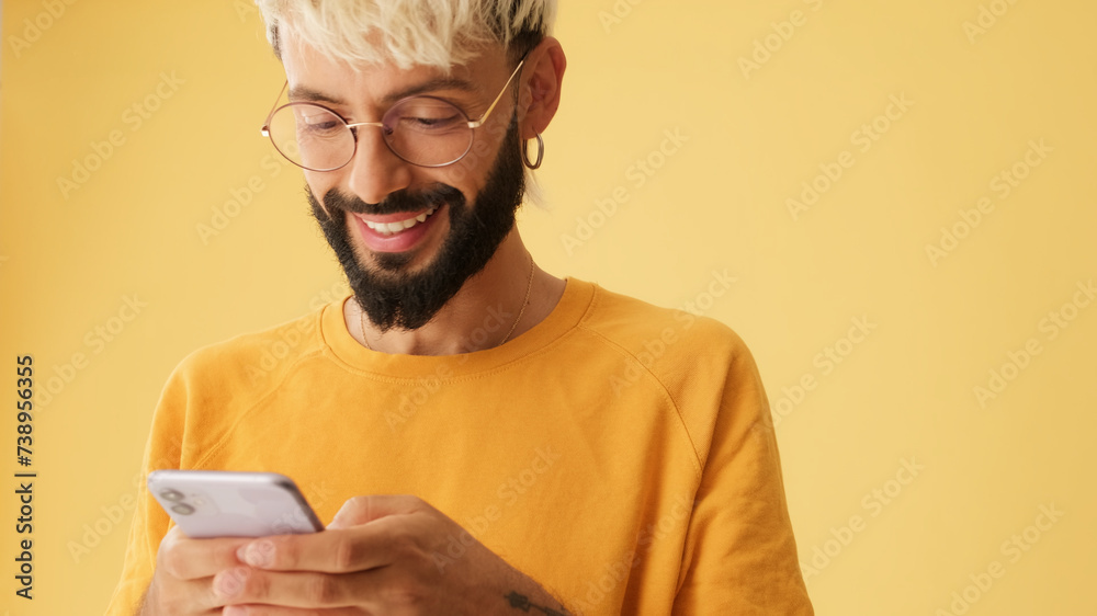 Guy with glasses, dressed in yellow T-shirt, laughs while reading sms, watching video on mobile phone, isolated on yellow background in studio Guy with glasses, dressed in yellow T-shirt, laughs while reading sms, watching video on mobile phone, isolated on yellow background in studio