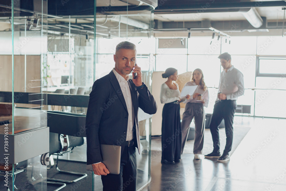 Focused businessman is talking phone with client while standing in office on colleagues background Focused businessman is talking phone with client while standing in office on colleagues background