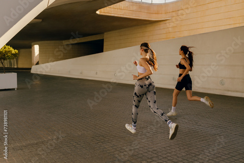 Two active women athlete running on sunny morning side by side on modern buildings background