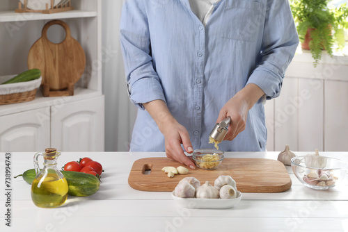 Woman squeezing garlic with press at white wooden table in kitchen, closeup