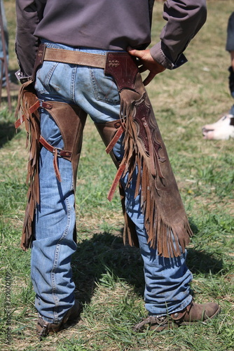 Rear view of an unrecognizable cowboy standing on green grass wearing brown leather chaps and denim jeans.