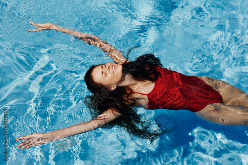 Happy woman swimming in the pool in red swimsuit with loose long hair relaxed in the sunshine, skin protection with sunscreen, concept of relaxing on vacation in tropical climate, lifestyle.