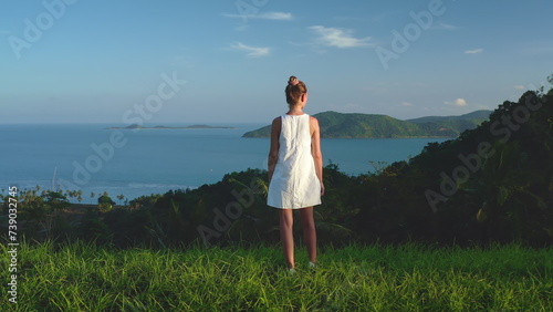 Woman enjoy fresh tropical island landscape standing on green grass hill looking on seascape horizon. Tourist female in white dress outdoor lifestyle travel on summer holiday vacation. Thailand, Samui photo