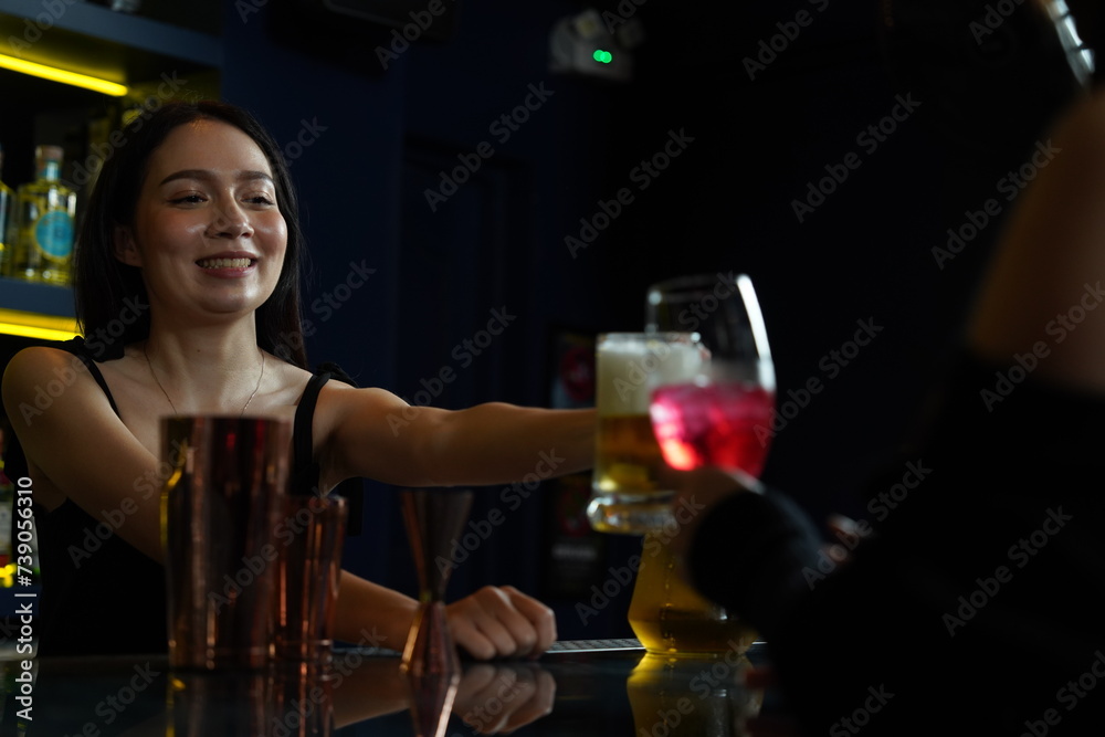 Asian female bartender holding a beer glass clinks with a female friend drinking red wine in a nightclub bar.