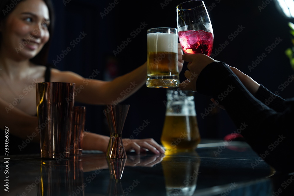 Asian female bartender holding a beer glass clinks with a female friend drinking red wine in a nightclub bar.