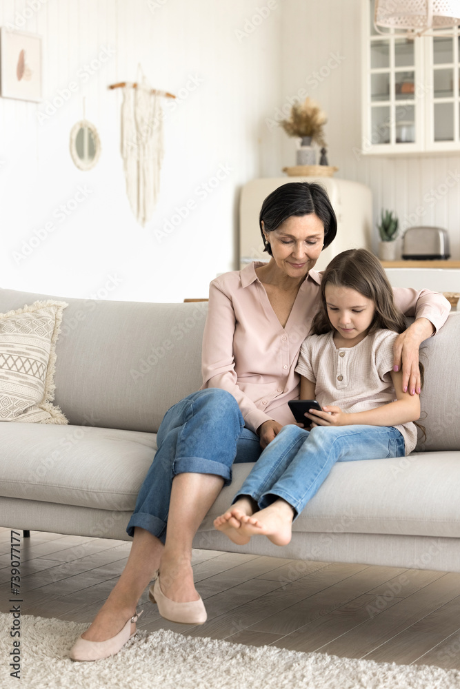 Preschooler girl spend time with grandmother using together modern smartphone, resting on couch enjoy new mobile application, web surfing internet, watching on-line videos, play new on-line videogame