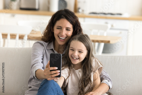 Happy Hispanic mother cuddling little 5s daughter, sit on sofa at home, taking selfie picture on smartphone camera, play online games, use funny mobile application, enjoy pastime leisure on internet
