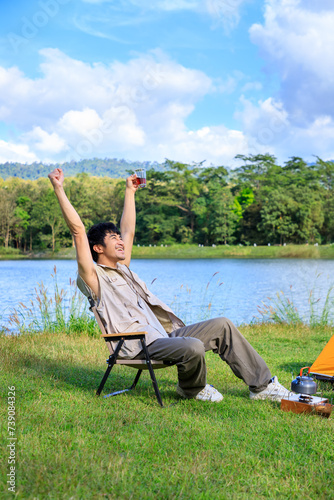 Asian male man sitting on chair, drinking coffee, feeling relax and raising hands, with camping tent beside lake and mountain.
