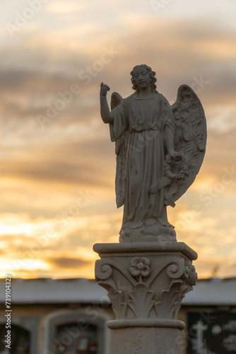  Silhouette of the statue of an angel, on a pedestal in the cemetery, against cloudy sky at dawn, backlit.