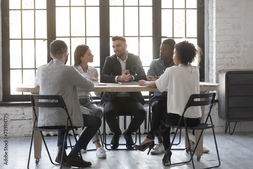 Happy male CEO holding meeting with employees at table in modern office, discussing work strategy. Competent, experienced professional sharing knowledge with office workers during business workshop