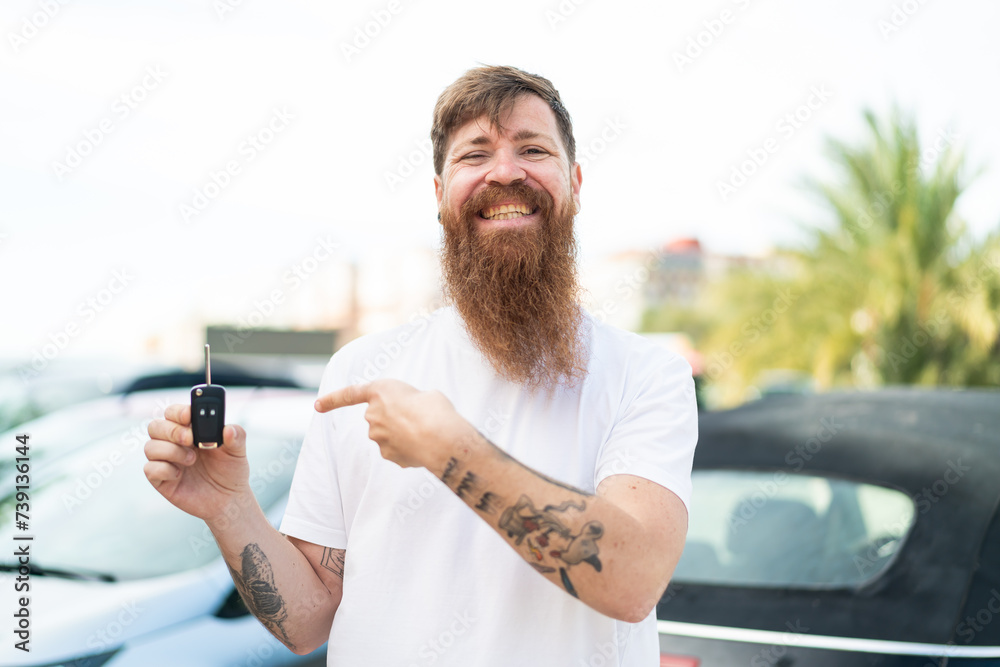 Redhead man with beard holding car keys at outdoors and pointing it Redhead man with beard holding car keys at outdoors and pointing it