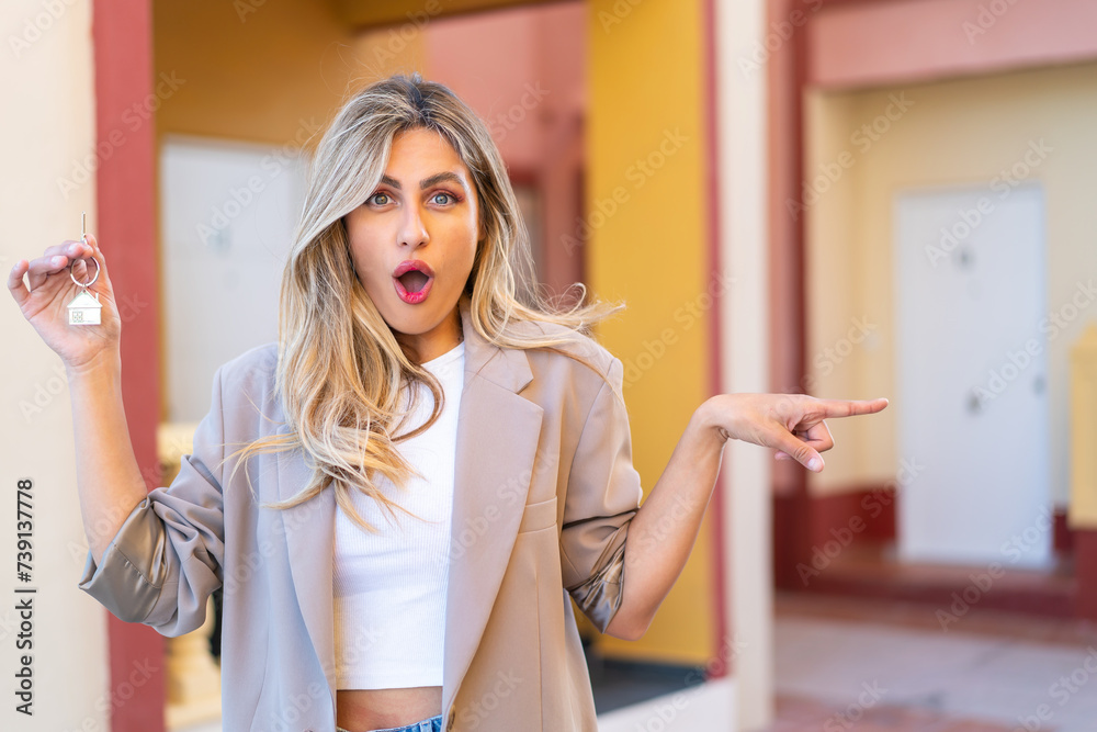 Pretty blonde Uruguayan woman holding home keys at outdoors surprised and pointing side Pretty blonde Uruguayan woman holding home keys at outdoors surprised and pointing side