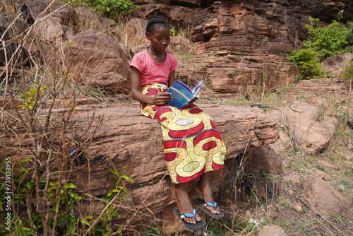 Young black elementary schoolgirl sitting on a rock reading in her textbook - Concept of female education and empowerment in Africa