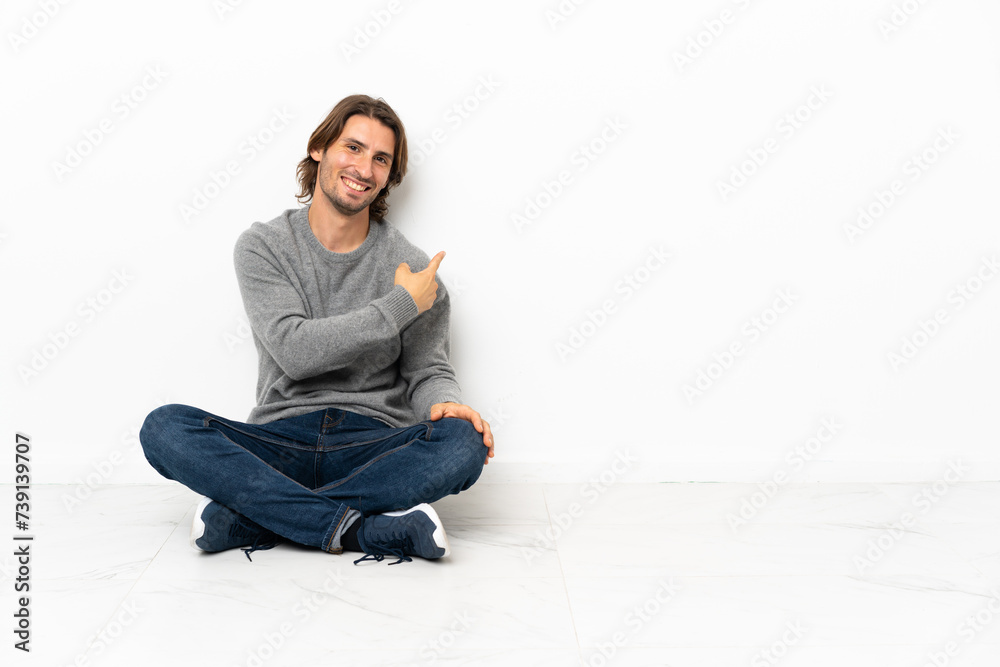 Young handsome man sitting on the floor over isolated background pointing back Young handsome man sitting on the floor over isolated background pointing back