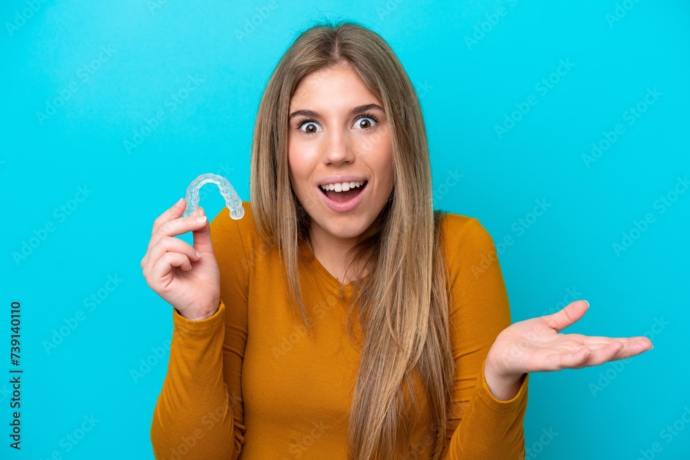 Young caucasian woman holding invisible braces isolated on blue background with shocked facial expression Young caucasian woman holding invisible braces isolated on blue background with shocked facial expression