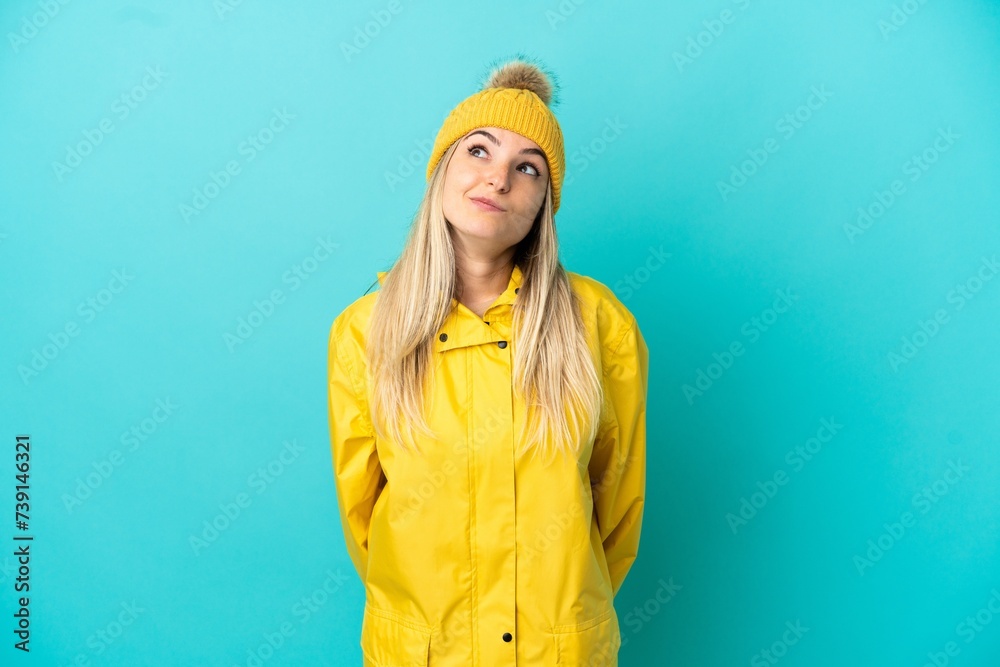 Young woman wearing a rainproof coat over isolated blue background and looking up Young woman wearing a rainproof coat over isolated blue background and looking up