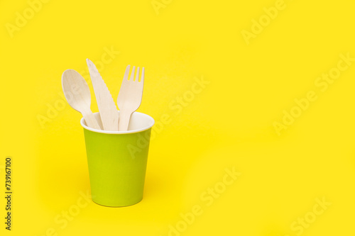 disposable wooden spoon  fork and knife in a green paper cup on a yellow background with copy space  recycled.